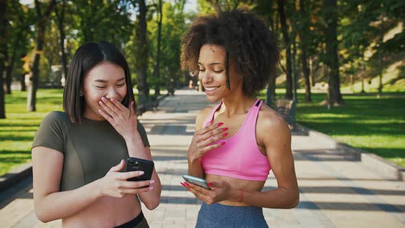 Outdoor Portrait of Asian and African American Female Friends Looking at Smartphone and Laughing in