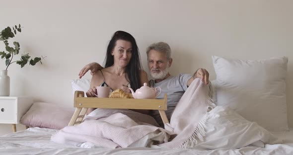 Wide Shot Portrait of Happy Senior Caucasian Man and Woman Lying in Bedroom with Tea and Croissants