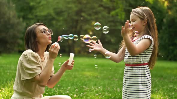 Beautiful Young Woman Sits on Green Grass in the Park and Blows Soap Bubbles