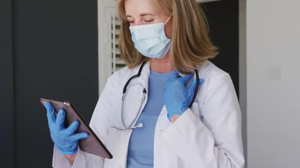 Caucasian senior female doctor wearing surgical gloves and face mask using tablet computer