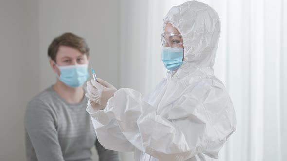 Concentrated Doctor or Nurse in Protective Suit and Eyeglasses Preparing Syringe with Vaccine As Man