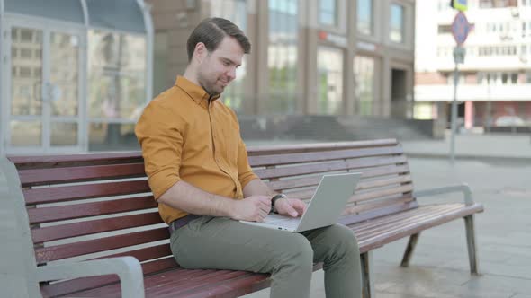 Young Man Celebrating Success on Laptop while Sitting Outdoor on Bench