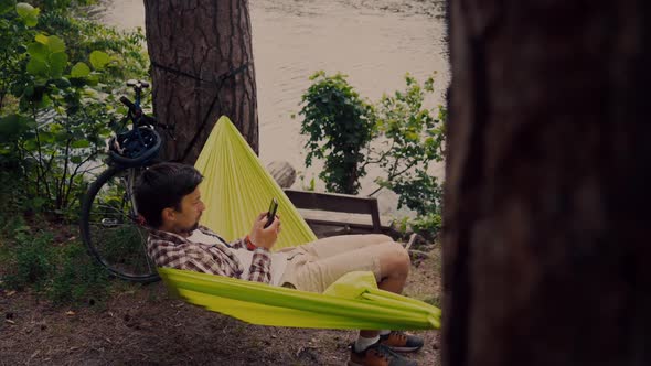 Man Cyclist is Resting Using the Phone in a Light Green Hammock on Nature Near the Lake After