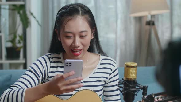 Woman Reading Comments On Smartphone And Speaking To Camera While Live Stream Playing Guitar At Home