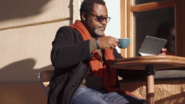 Positive African man in eyeglasses leafing tape by tablet and drinking coffee