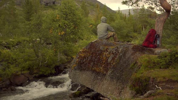 A Traveler with a Red Backpack Sitting on the Rock Above the Mountain Waterfall