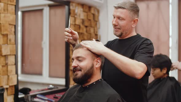 An Adult Caucasian Barber with Gray Beard Cuts the Hair of Hipster with a Beard While Chatting at