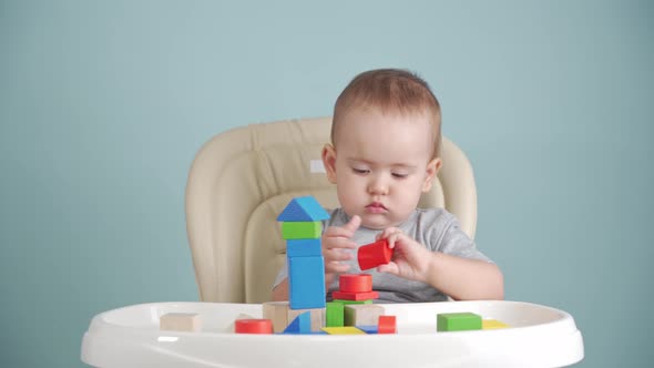 A joyful newborn 12-17 months old builds a tower of wooden cubes sitting on a chair.