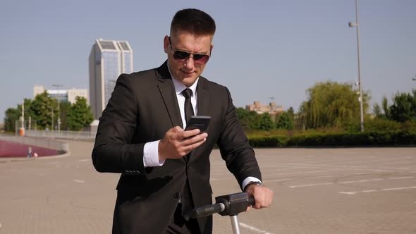 A Businessman in Suit with a Phone in the Park Next To His Electric Scooter