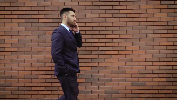 Businessman Dressed in Shirt Talking Smartphone While Walks on the Background of a Modern Brick Wall