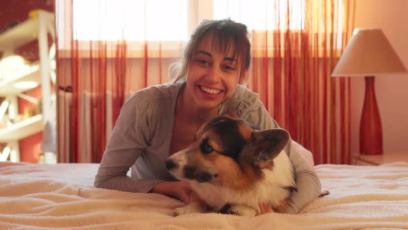 Close-up Portrait of Pleased Woman Lying on Bed at Home with Her Loved Dog Welsh Corgi.