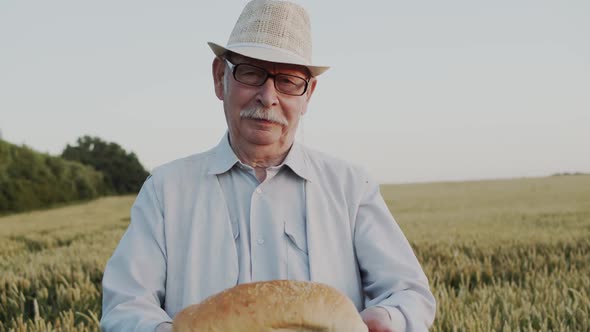 A Mustachioed Old Man with Glasses in Hat Presents a Loaf of Bread To the Camera