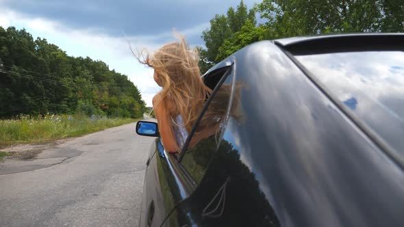 Small Girl Leaning Out of Car Window While Riding Through Country Road at Overcast Weather. Little
