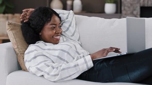 Smiling African Woman Student Girl Freelancer User Lying on Couch Using Laptop Typing Message Online