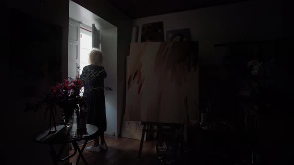 A Middle Aged Woman Artist in a Dark Room of the Gallery Opens a Window