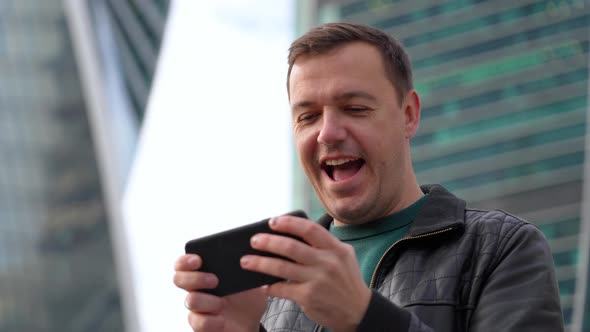 Emotional Millennial Guy Playing Video Game on Smartphone in Front of Modern Buildings