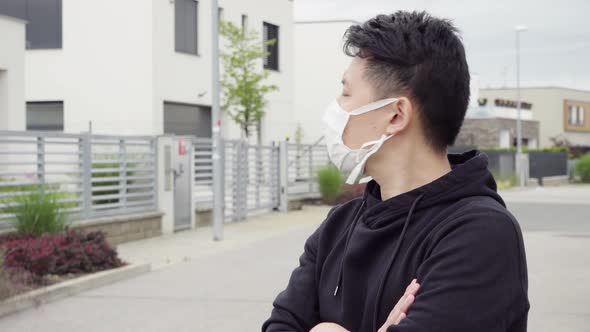 A Young Asian Man in a Face Mask Looks Around, Thoughtful, in an Empty Suburban Area