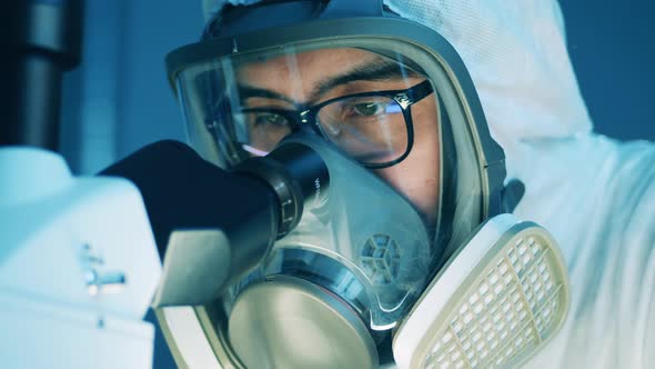 Male Specialist in a Hazmat Mask is Looking Into Microscope