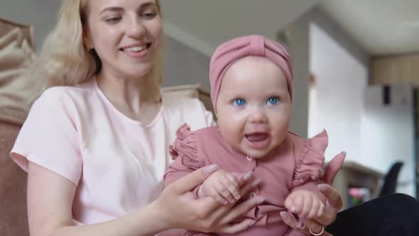 Baby Girl with Blond Hair and Blue Eyes in Pink Clothes Sits in Her Mother's Arms and Actively Moves