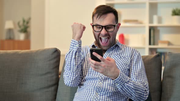 Yong Man Celebrating Success on Smartphone at Home