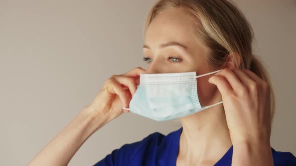 Closeup Shot of a Caucasian Blonde Female Nurse in a Blue Uniform Putting on a Protective Face Mask