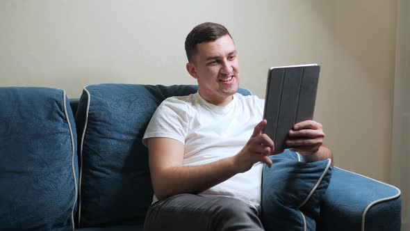 Cheerful Young Guy Smiling and Greeting Friend While Sitting on Blue Sofa and Using Video Chat App