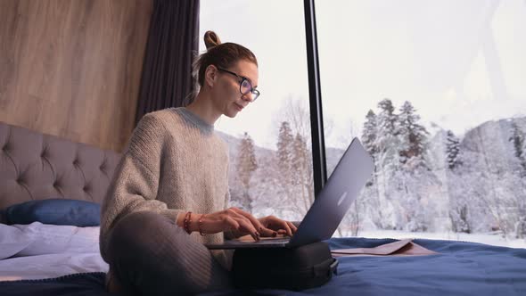 A Woman Freelancer with Glasses Sits on a Bed in a Country House with Panoramic Windows Behind Which