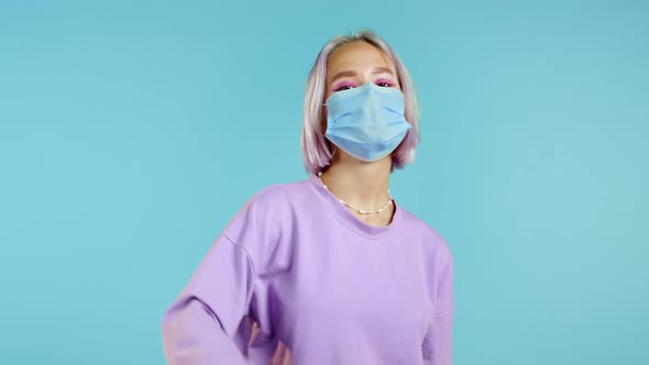 Cute Woman with Medical Mask and Violet Dyed Hairstyle Showing Thumb Up Sign Over Blue Background