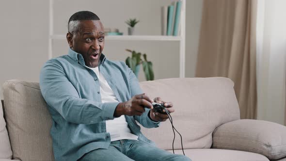 Excited Worried Emotional Adult African American Male Gamer Sitting on Sofa in Living Room Playing