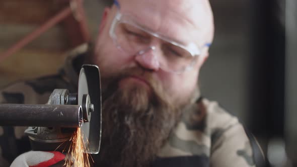 Male Construction Worker in Protective Glasses Works By Cutting Metal Pipe with an Angle Grinder