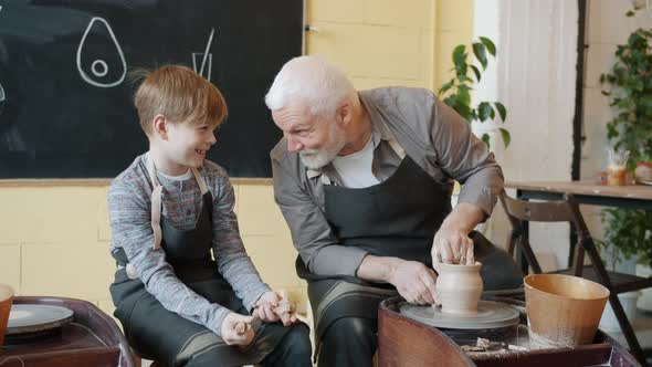 Grandfather Potter Making Vase on Throwing-wheel and Talking To Cheerful Kid in Studio