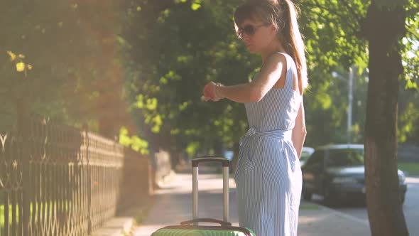 Young Woman Walking on City Street Sidewalk with Green Suitcase on Summer Day