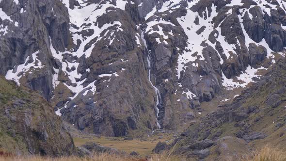 Static, hiker walks past distant waterfall in snow capped landscape, Routeburn Track New Zealand