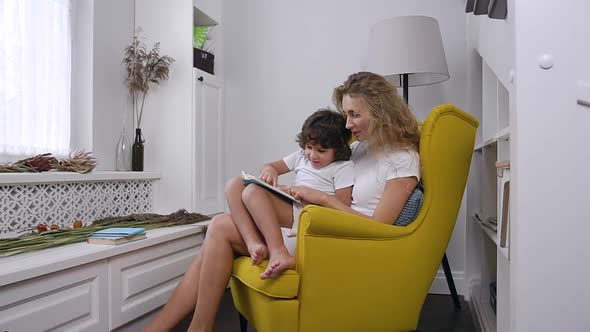 Mom Reads Child book to her little son while sitting in comfortable yellow armchair