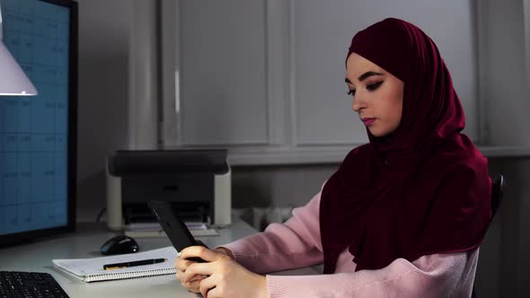 Portrait of Young Woman in Hijab Searching Information in a Smartphone and Making Notes in Copybook