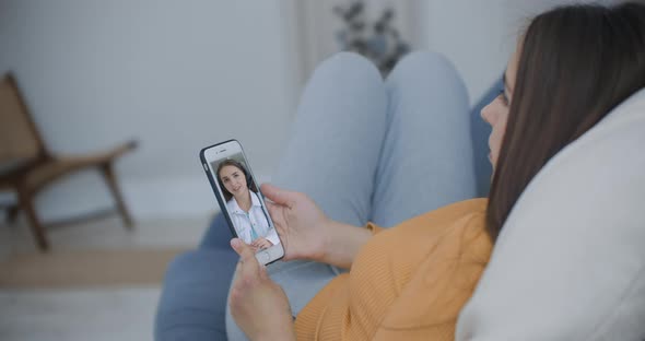 A Woman Lying on the Couch Talking To a Woman Doctor on Video Using a Smartphone. Remote Medical