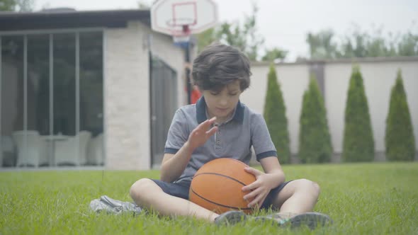 Thoughtful Caucasian Boy Sitting on Green Grass with Ball As Blurred Brother Climbing on Basketball