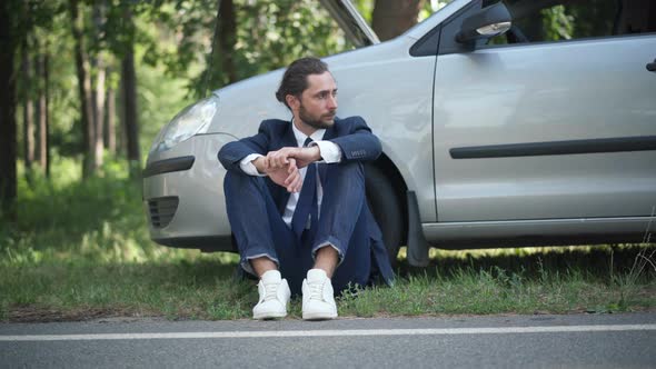 Front View Portrait of Stressed Male Driver Holding Head in Hands Sitting at Broken Car with Open