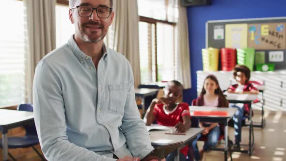 Portrait of happy caucasian male teacher in classroom with children looking at camera