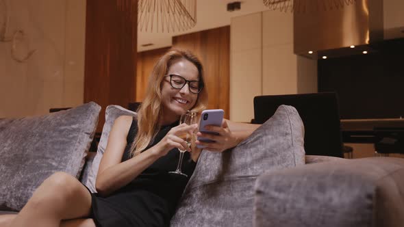 Woman in Evening Dress with Wineglass Laughs on the Sofa with Phone