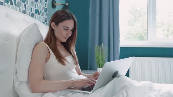 Calm Woman Sitting Up in Her Hotel Bed Balances Her Laptop on Her Legs As She Taps on the Keyboard