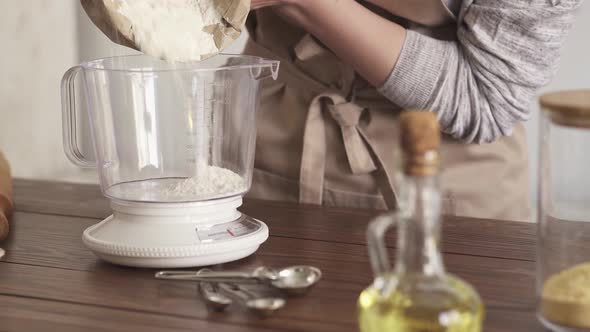 Weighing Flour On A Scale. Female Hands Pour Flour From A Paper Bag Into A Bowl Of Kitchen Scales