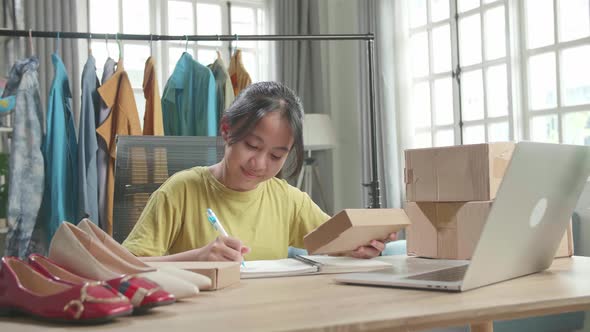 Young Girl Online Seller Looking At The Package And Writing In The Notebook While Selling Clothes