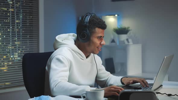 Black Gamer in White Hoodie and with Headphones Playing Games on Computer in the Evening