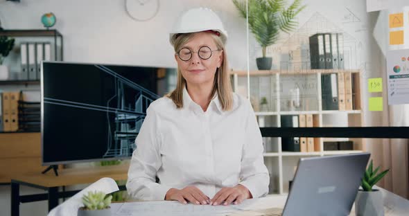 Female Architect in Helmet Sitting at Workplace in front of Camera in Modern Design Office