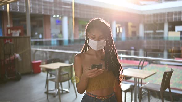 Young black woman in face mask uses smart phone in commercial mall