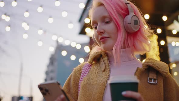 Young Cheerful Pink Haired Woman Wearing Headphones Chatting on Smartphone Enjoying Takeout Coffee