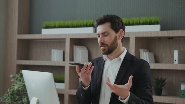 Businessman Seated at Workplace Desk Holding Smart Phone Speaking Through Microphone Sending Voice