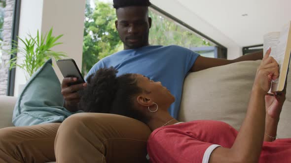 Video of happy african american couple lying on sofa with smartphone and book