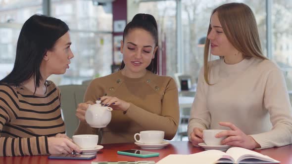 Young Brunette Caucasian Woman Pouring Tea Into Cups and Talking with Female Friends. Three